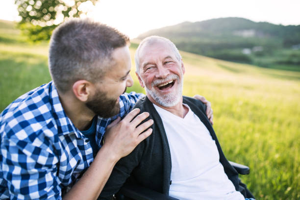 un hijo adulto hipster con senior padre en silla de ruedas en una caminata en la naturaleza en el ocaso, riendo. - walking senior adult family old fotografías e imágenes de stock