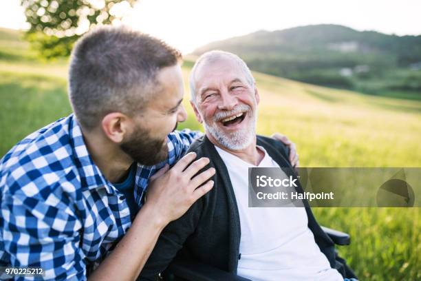 Photo libre de droit de Un Fils Adulte Hipster Avec Père Senior En Fauteuil Roulant Sur Une Promenade Dans La Nature Au Coucher Du Soleil En Riant banque d'images et plus d'images libres de droit de Troisième âge