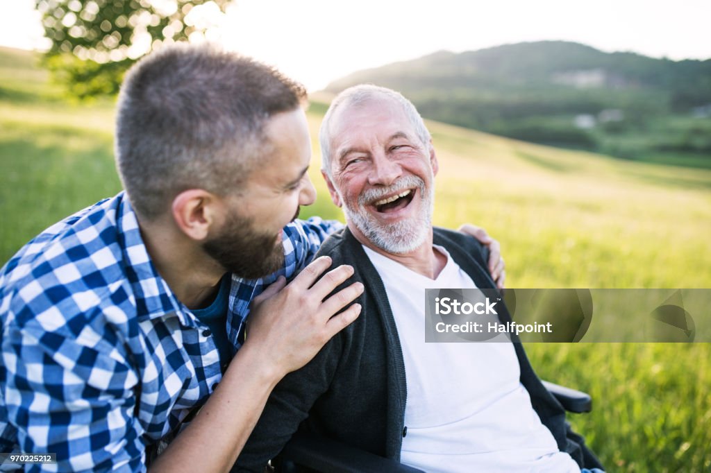 An adult hipster son with senior father in wheelchair on a walk in nature at sunset, laughing. An adult hipster son with his senior father in wheelchair on a walk on a meadow in nature at sunset, laughing. Close up. Senior Adult Stock Photo