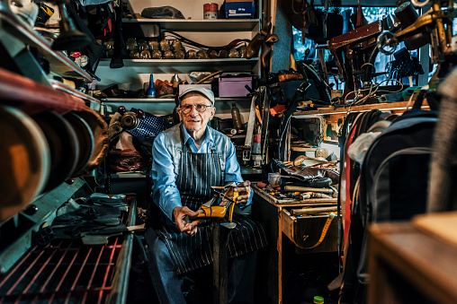 Photo of senior craftsman wearing hat and eyeglasses making luxury handmade man or woman shoes in his small workshop while looking at camera. Professional diligent old shoemaker heeling footwear on machine at the shop.