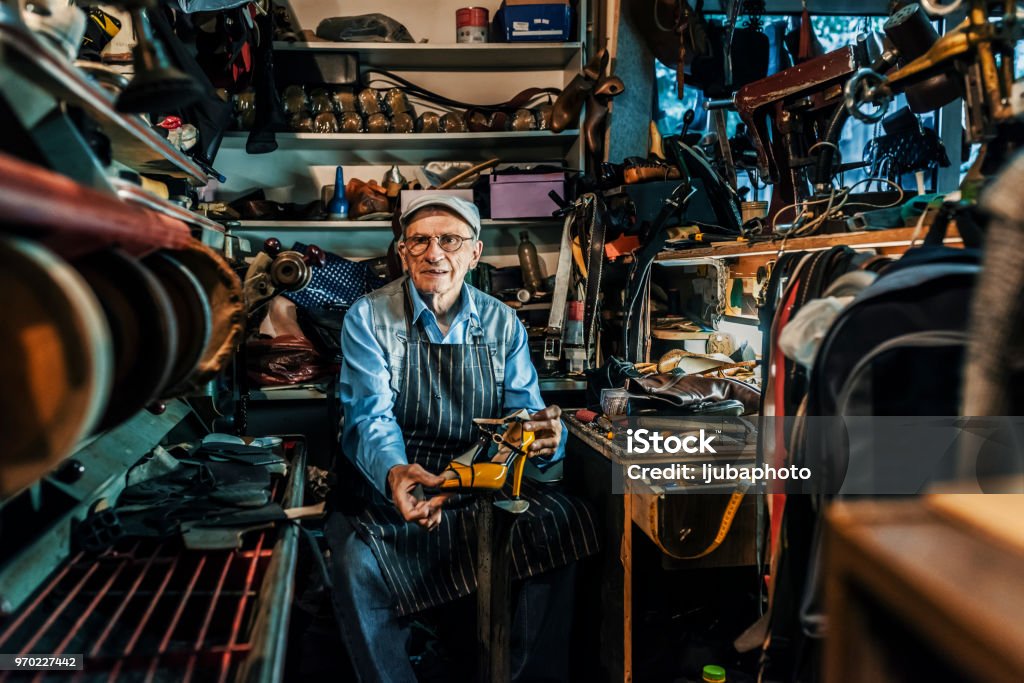 He had an eye for detail Photo of senior craftsman wearing hat and eyeglasses making luxury handmade man or woman shoes in his small workshop while looking at camera. Professional diligent old shoemaker heeling footwear on machine at the shop. Craftsperson Stock Photo