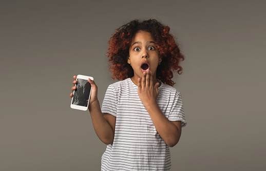 Little african-american girl with broken smartphone at gray studio background, copy space