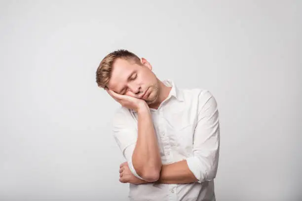 Sleepy young caucasian man in white shirt yawning, closing his mouth with hand, isolated on gray background. Concept of overworked guy