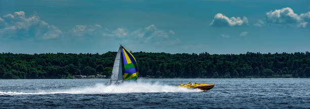 Panorama of a speedboat leaving a roostertail while passing a sailboat with a cloudy blue sky and distant shoreline. Panorama of a speedboat leaving a roostertail while passing a sailboat with a cloudy blue sky and distant shoreline. racing boat photos stock pictures, royalty-free photos & images