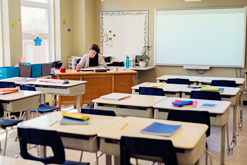 Elementary teacher, working at her desk in an empty classroom. She is wearing a light sweater and her brown hair is tied in a ponytail. Lots of empty desk in front of her. Electronic board at her side. Horizontal indoors full length shot with copy space.