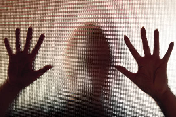 Spooky silhouette of trapped abused woman with hands pressed against glass window Colour backlit image of the silhouette of a woman with her hands pressed against a glass window. The silhouette is distorted, and the arms elongated, giving an alien-like quality. The image is sinister and foreboding, with an element of horror. It is as if the 'woman' is trying to escape from behind the glass. Horizontal image with copy space. Trafficking stock pictures, royalty-free photos & images