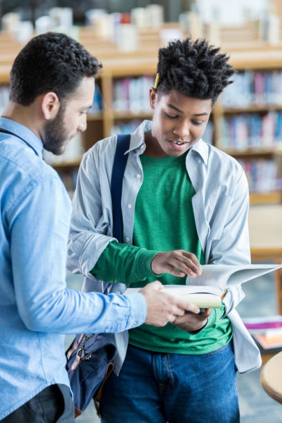 Male high school student asks teacher question A male high school student stands in his school library and flips through a book with his teacher.  He is asking a question. guru photos stock pictures, royalty-free photos & images