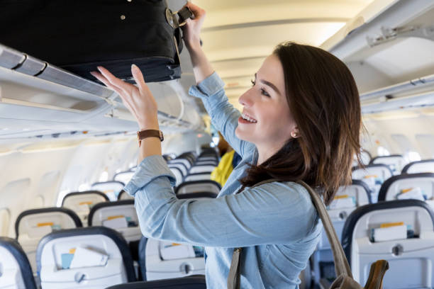 Young woman puts carry on bag in overhead bin of airliner A cheerful young woman stands in the aisle of a commercial airliner and pushes her suitcase into an overhead bin. hand luggage stock pictures, royalty-free photos & images