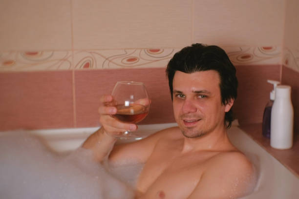 istock Young man takes a bath with foam and going to drinks whiskey. Smiling and looking at camera. 970361436