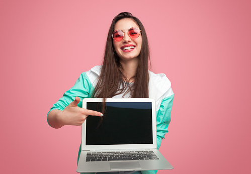 Trendy bright girl in pink sunglasses holding laptop and pointing at screen smiling at camera on pink