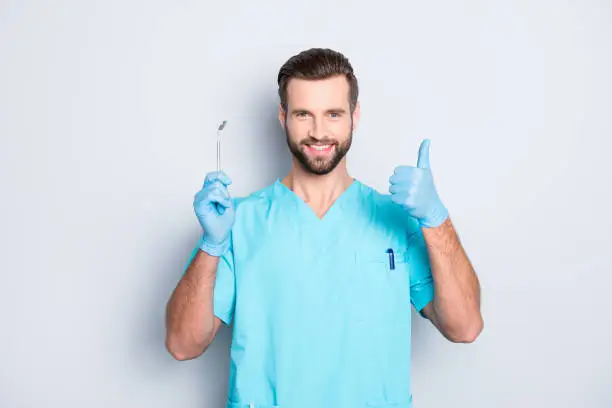 Portrait of joyful cheerful dental assistant in lab blue uniform gesturing thumbup approve advice yes done sign with finger, having tool in hand, isolated on grey background