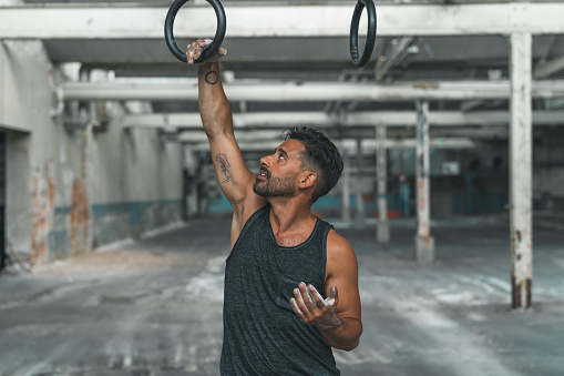 A muscular man in a tank top is exercising with gymnastic rings inside an abandoned warehouse.