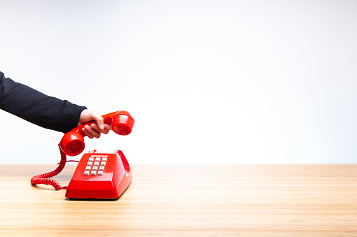 Business woman hanging up old red telephone, classic red telephone receiver in hand wearing black suit, old telephone isolated on white background and wooden desktop.