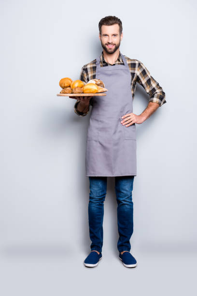 полноразмерный портрет тела положительного привлекательного пекаря в джинсах, обуви, рубашке, фартуке с щетиной, имеющих поднос с хлебобул - whole meal bread стоковые фото и изображения