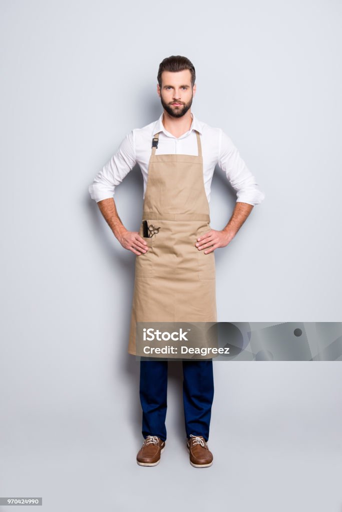 Full size fullbody portrait of virile harsh barber in uniform holding arms on waist, looking at camera, isolated over grey background Apron Stock Photo