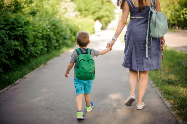 vista posterior de la madre caminando por la calle con un hijo pequeño con una mochila en día soleado - greenback fotografías e imágenes de stock