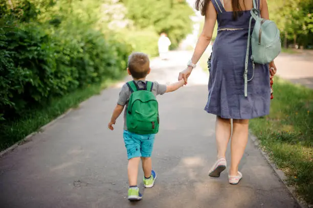 Photo of Back view of mother walking down the street with a little son with a backpack on sunny day