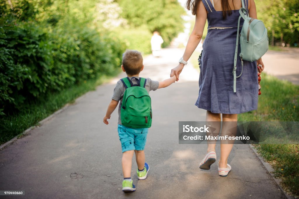 Vista traseira da mãe andando pela rua com um filho pequeno com uma mochila em dia ensolarado - Foto de stock de Criança royalty-free