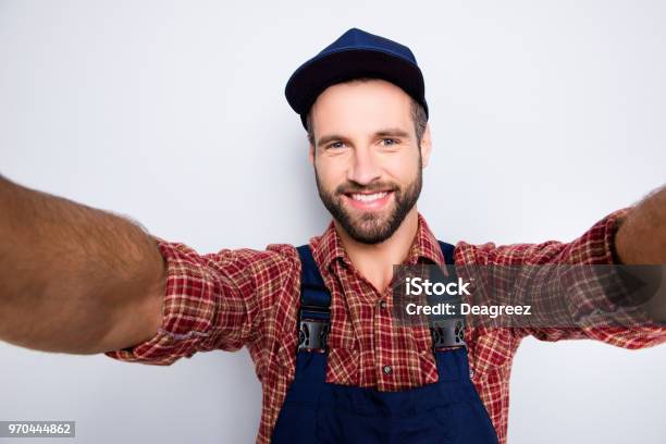 Self Portrait Of Joyful Cheerful Mechanic With Stubble In Blue Overall Shirt Shooting Selfie On Front Camera With Two Hands Isolated On Grey Background Stock Photo - Download Image Now