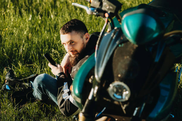 istock Young caucasian man sitting on the grass leaning on his bike and using a smartphone while looking into camera smiling in his traveling time while resting near the road. 970489908