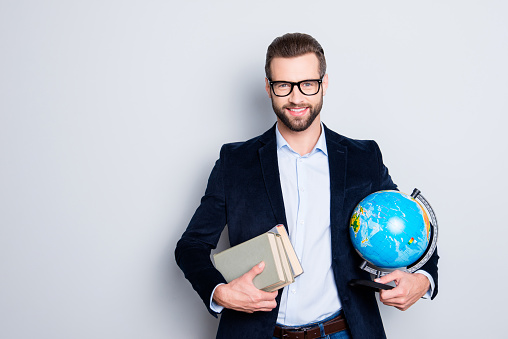 Portrait of stylish joyful teacher in shirt jacket with stubble having books and round globe, earth in hands, looking at camera, isolated on grey background