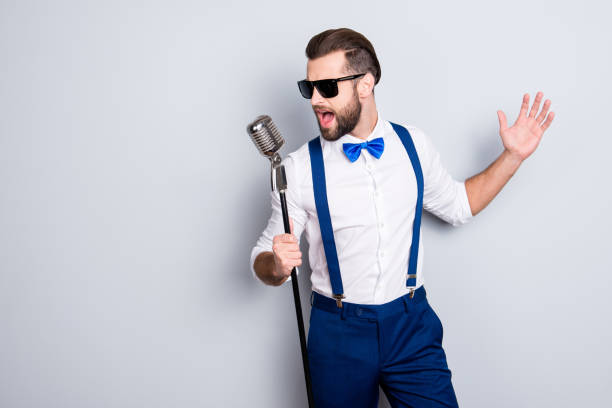 Portrait of handsome attractive singer in blue pants with suspenders and black glasses, singing hit with open mouth in microphone gesture with hand isolated on grey background Portrait of handsome attractive singer in blue pants with suspenders and black glasses, singing hit with open mouth in microphone gesture with hand isolated on grey background solo performance stock pictures, royalty-free photos & images