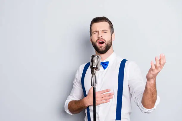 Photo of Portrait of talented attractive singer with bristle stubble in elegant classic outfit, singing hit with open mouth in microphone gesture with hand isolated on grey background