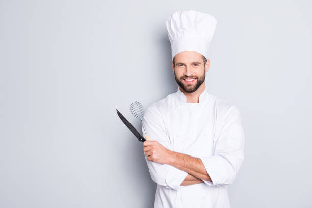 ritratto di chef dentato positivo cucinare in berretto, abito bianco con stoppie con strumenti a braccia incrociate guardando la fotocamera isolata su sfondo grigio - copricapo abbigliamento foto e immagini stock