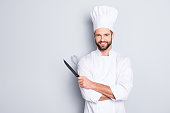 Portrait of positive toothy chef cook in beret, white outfit with stubble having tools in crossed arms looking at camera isolated on grey background