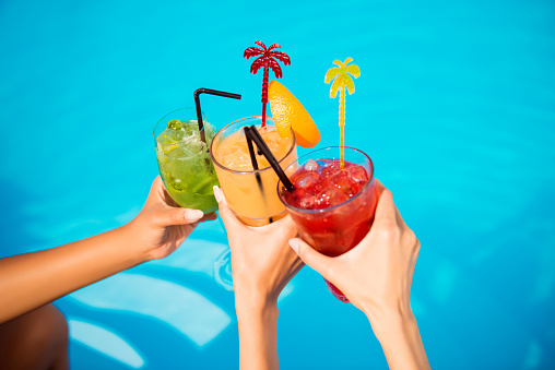 Three ladies cheering with tasty nectars with ice cubes, with black straws, palms decoration, in swim pool, transparent clean clear blue water, sun shines, smooth tanned skin, carefree festive mode