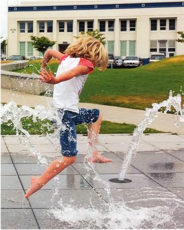 Photograph of a young blond girl leaping over sprays of water on a summer day