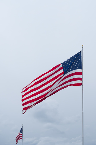 Two American flags with stars and stripes blowing in a breeze against cloudy gray sky.
