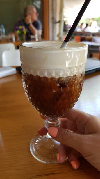Cold cappuccino with whipped cream topping in vintage glass holding in female hand with summer nails. Greek style freddo cappuccino with thick milk cream and ice cubes Summer drink from first person point of view. freddo cappuccino stock pictures, royalty-free photos & images