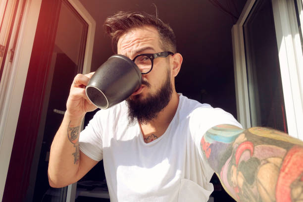 Man with tattoos making window selfie and drinking morning coffee