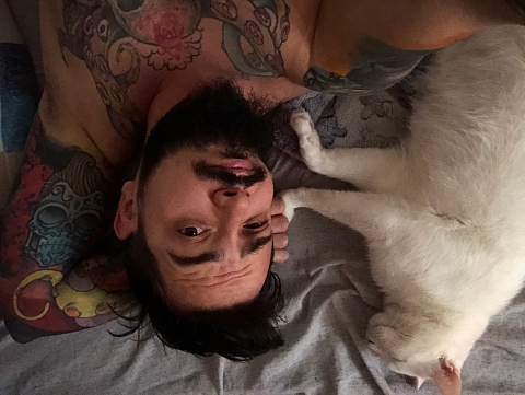 Man with tattoos laying with white cat in bed at home and making selfie.