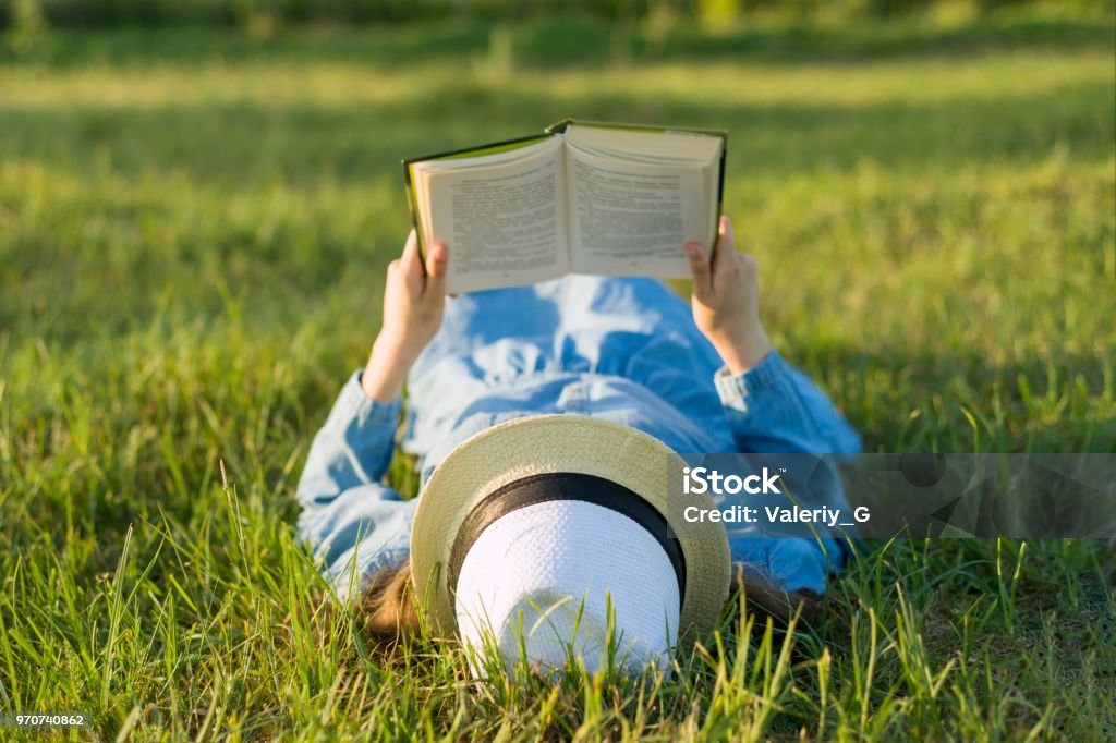 Ragazza in abito e cappello giace su un libro di lettura in erba verde. Vista dall'alto - Foto stock royalty-free di Leggere