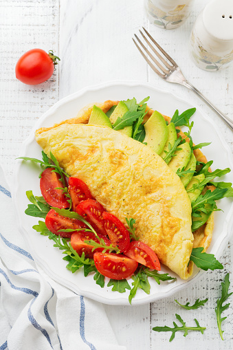Omelette with avocado, tomatoes and arugula on white ceramic plate on light stone background. Healthy breakfast. Selective focus. Top view. Copy space.