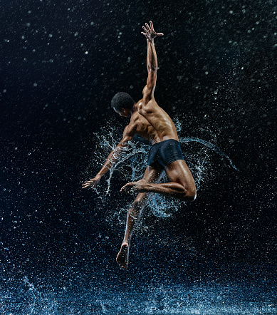 Athletic afro american ballet dancer dancing with water drops over blue background. Aqua, spray, splash, rain concept. Modern contemporary choreography with young professional man
