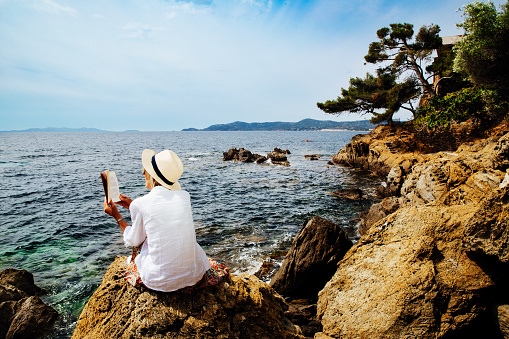 Senior woman reading a book on a rocky seashore in Provence, France.