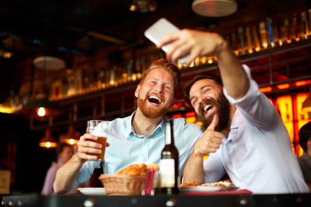Male friends in the bar posing for a selfie Bearded male couple of friends having a good time in the pub with beer drinks as one of them is holding mobile phone and capturing a selfie image. alcohol abuse photos stock pictures, royalty-free photos & images