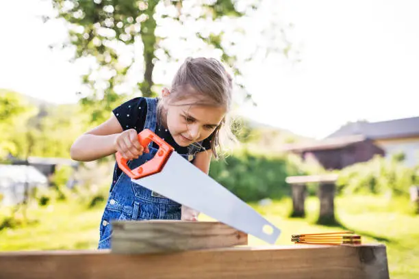 Photo of A small girl with a saw outside, making a wooden birdhouse.