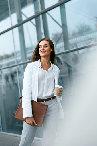 Beautiful Woman Going To Work With Coffee Walking Near Office Building. Portrait Of Successful Business Woman Holding Cup Of Hot Drink In Hand On Her Way To Work On City Street. High Resolution.