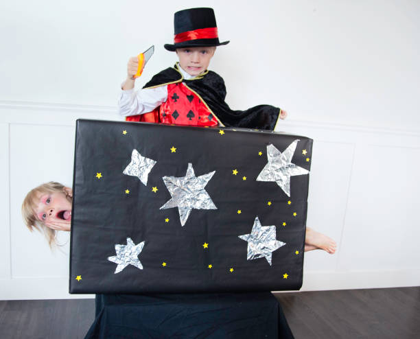 Young Magician saws  his Pretty Assistant Brother and Sister play magician and assistant, while brother pretends to cut through box sister is laying in magic show stock pictures, royalty-free photos & images