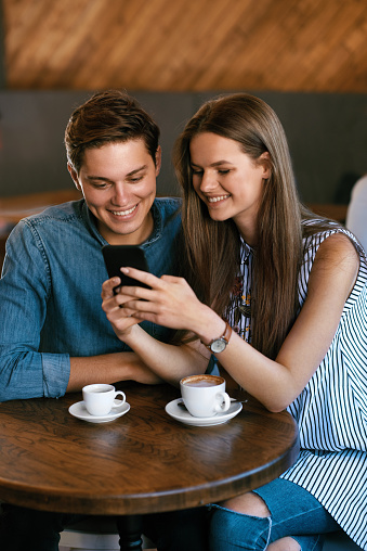 Beautiful Couple In Cafe Using Phone. Beautiful Smiling Woman And Handsome Man Drinking Coffee, Using Mobile Phone While Spending Time In Coffee Shop. High Quality Image.