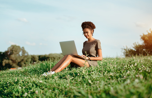 Cheerful young African-American female student with the laptop on the lawn outdoors on a summer day; smiling Brazilian undergraduate girl is making homework via the netbook while sitting on the meadow