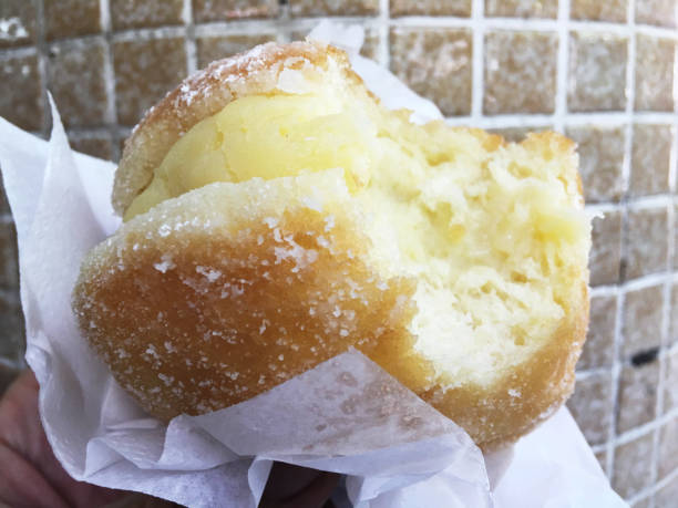 Hand holding fresh Brazilian sonho sugar-coated doughnut with a bite taken out exposing custard cream filling