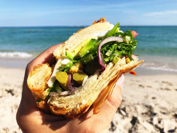 Hand holding gourmet vegetarian croissant sandwich on a bright beach background
