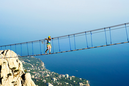 Young woman walking on hanging rope bridge at panoramic sea landscape. Leisure activity and extreme sport