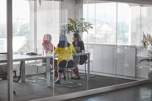 Full length of businesswomen discussing at board room. Female colleagues are seen through glass wall. They are working at office.