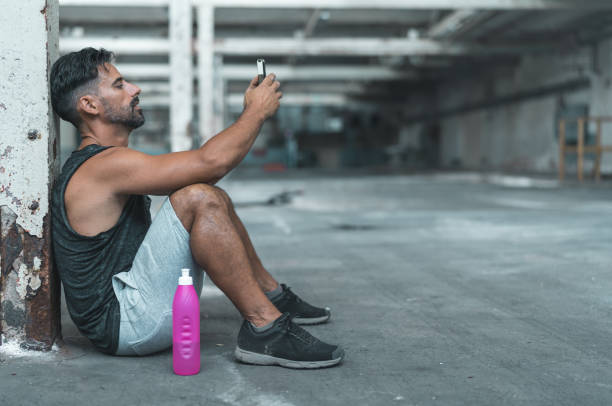 A muscular man using a mobile smart phone while exercising inside an abandoned warehouse.